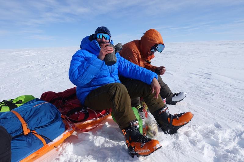 To menn på en pulk på grønlandsisen. EN ser i kamera og holder en flaske.